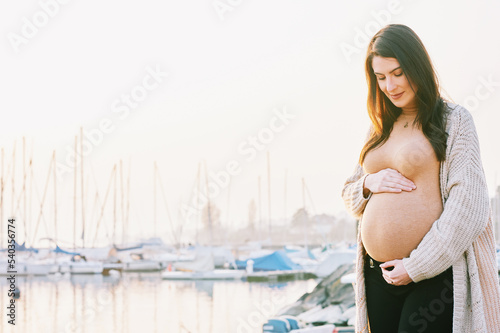 Fototapeta Naklejka Na Ścianę i Meble -  Outdoor portrait of beautiful pregnant woman touching belly, early morning walk next to lake