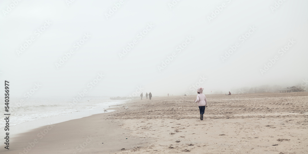 young girl is playing on an empty beach during a foggy, cold day