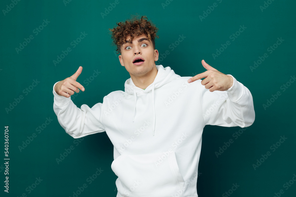 a cute, funny, emotional man stands on a green background in a white hoodie and looks at the camera with his mouth wide open, pointing his fingers at his clothes. Horizontal Studio Photography