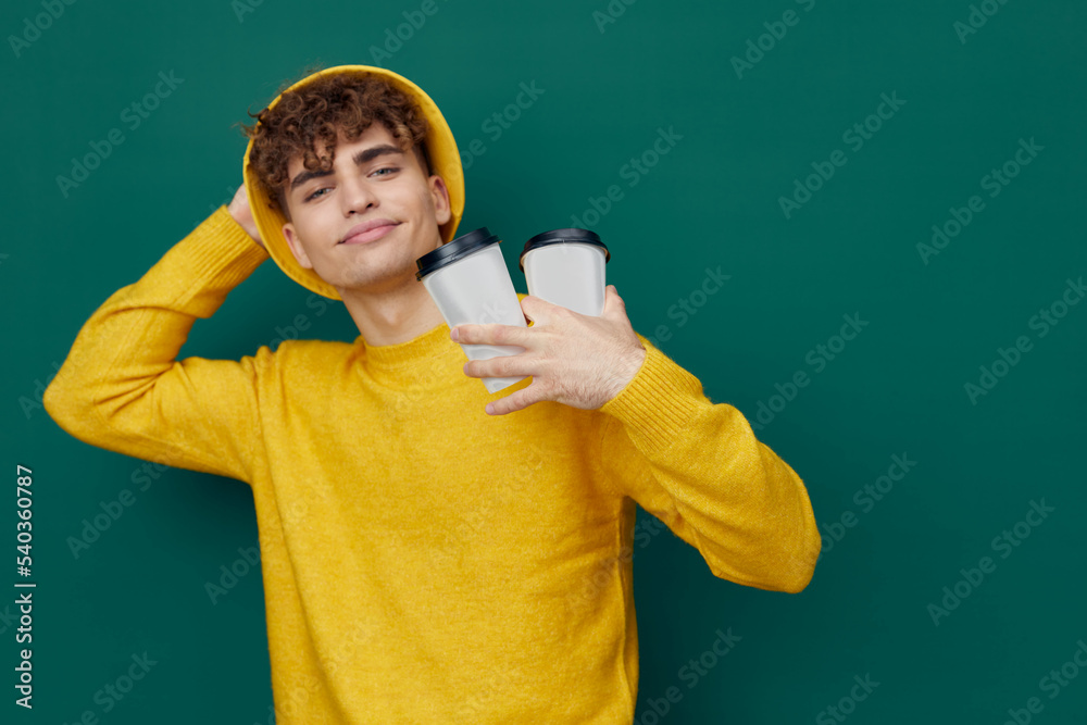  man stands on a green background in a yellow T-shirt and hat holding two cardboard glasses in his hands, and holding his head with the other hand. Horizontal photo for inserting an advertising layout