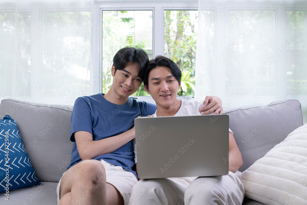 Young Asian gay couple communicating online on laptop. LGBT male couple embracing watching movie webinar on laptop studying e-learning together, doing paperwork, paying bills online in home. Young Asian gay couple communicating online on laptop. LGBT male couple embracing watching movie webinar on laptop studying e-learning together, doing paperwork, paying bills online in home.