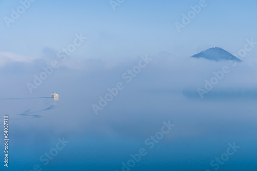 幻想的な霧の中に浮かび上がる山と湖を進む白い船 © SP studio