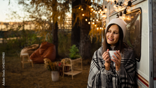Caucasian woman in a knitted hat wrapped in a plaid and drinks a warming drink outdoors. Travel in a motor home in the fall. © Михаил Решетников