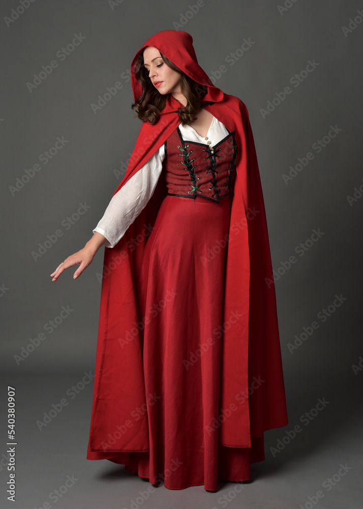 Full length portrait of beautiful brunette woman wearing red medieval fantasy costume with long skirt and flowing hooded cloak.
Standing pose with gestural hand poses, isolated on grey studio backgrou