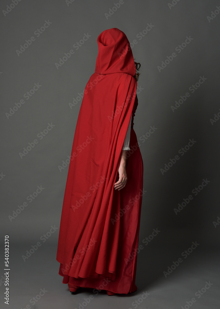 Full length portrait of woman wearing red medieval fantasy costume, flowing hooded cloak. Standing pose in backview, gestural hand poses, walking away from camera isolated on grey studio background. Full length portrait of woman wearing red medieval fantasy costume, flowing hooded cloak. Standing pose in backview, gestural hand poses, walking away from camera isolated on grey studio background.