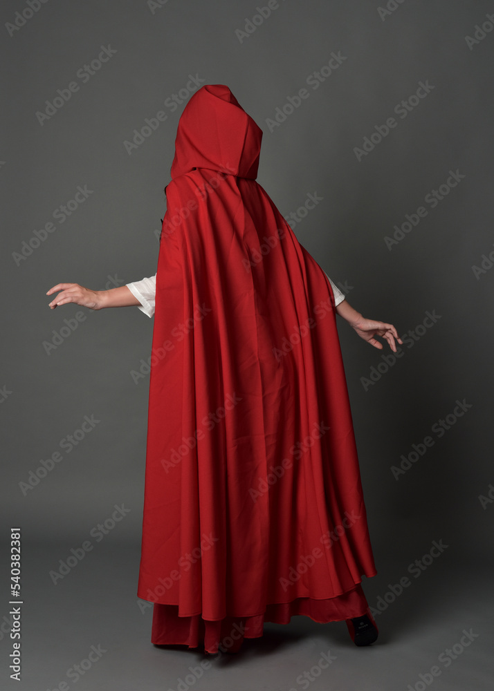 Full length portrait of woman wearing red medieval fantasy costume, flowing hooded cloak. Standing pose in backview, gestural hand poses, walking away from camera isolated on grey studio background.