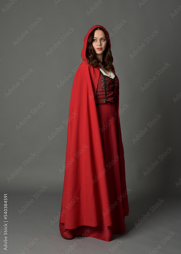 Full length portrait of woman wearing red medieval fantasy costume, flowing hooded cloak. Standing pose side profile, gestural hand poses, walking away from camera isolated on grey studio background.