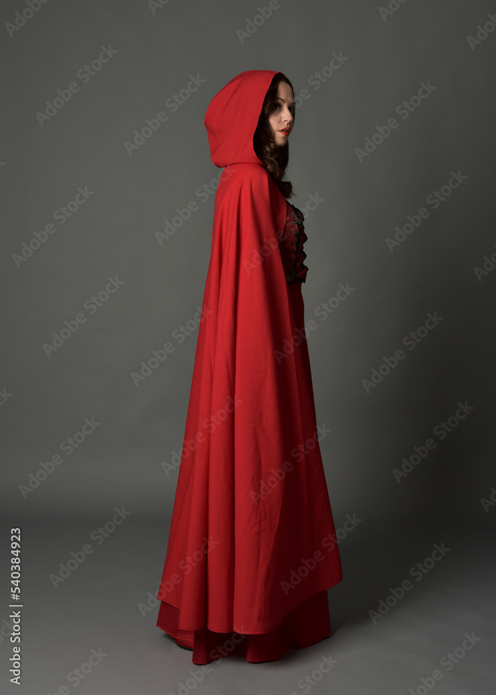 Full length portrait of woman wearing red medieval fantasy costume, flowing hooded cloak. Standing pose side profile, gestural hand poses, walking away from camera isolated on grey studio background.