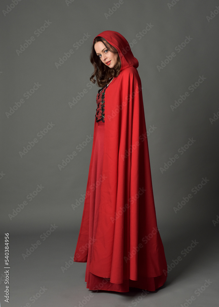 Full length portrait of woman wearing red medieval fantasy costume, flowing hooded cloak. Standing pose side profile, gestural hand poses, walking away from camera isolated on grey studio background. Full length portrait of woman wearing red medieval fantasy costume, flowing hooded cloak. Standing pose side profile, gestural hand poses, walking away from camera isolated on grey studio background.