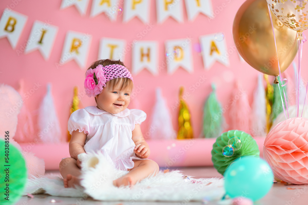 Smiling baby girl sits near paper balls and balloons in holiday dress on pink birthday background