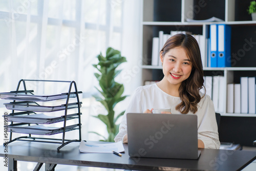 Beautiful Asian businesswoman working on a desk with a laptop with a smiling face while looking at the document received in the office.