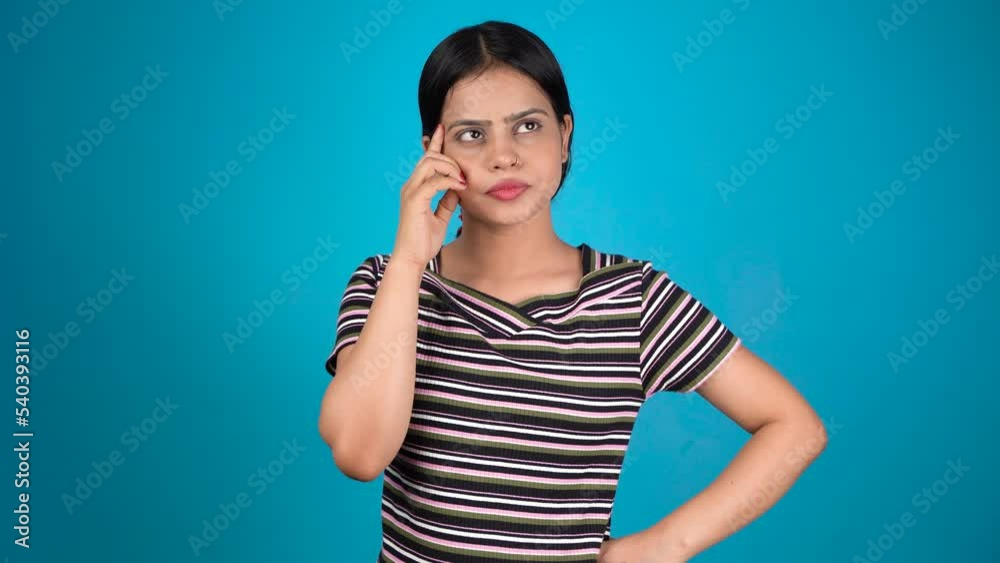 Pretty young woman looking up empty space deep thinking, holding arm on chin, isolated blue color background Pretty young woman looking up empty space deep thinking, holding arm on chin, isolated blue color background