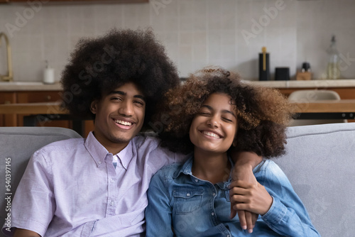 Lovely African teenager, smiling girl and guy sit on sofa look at camera, spend time together at home. Portrait of friendly roommates in good relations, first romantic relationships, couple in love