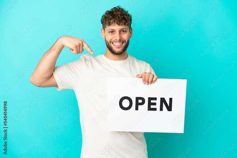 Young handsome caucasian man isolated on blue background holding a placard with text OPEN and pointing it Young handsome caucasian man isolated on blue background holding a placard with text OPEN and pointing it