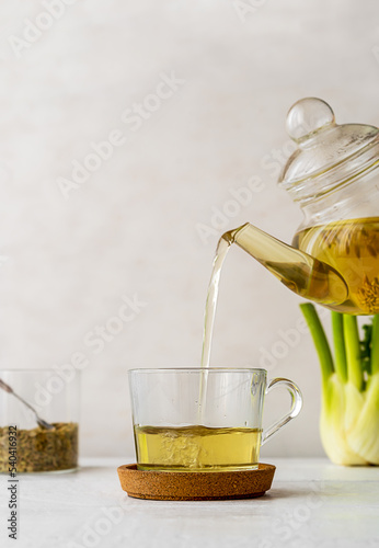 Fennel tea in a glass cup, fresh fennel bulb, seeds and tea pot pouring tea to cup. White wooden table with light background