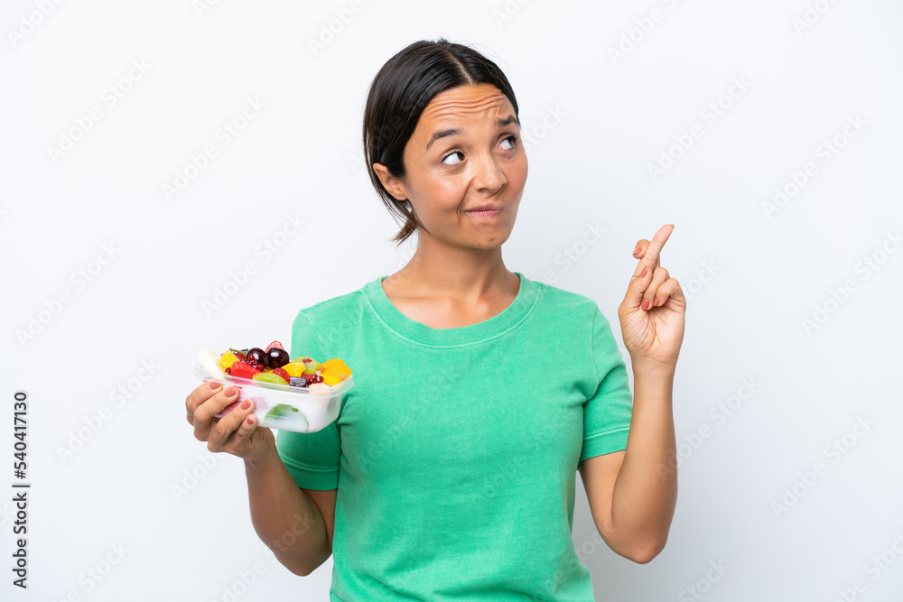 Young hispanic woman holding a bowl of fruit isolated on white background with fingers crossing and wishing the best Young hispanic woman holding a bowl of fruit isolated on white background with fingers crossing and wishing the best