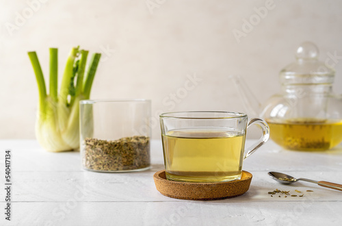 Fennel tea in a glass cup, fresh fennel bulb, seeds in jar, tea pot on white wooden table with light background