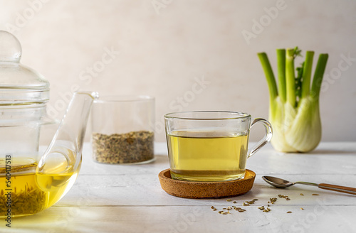 Fennel tea in a glass cup, fresh fennel bulb, seeds in jar, tea pot on white wooden table with light background