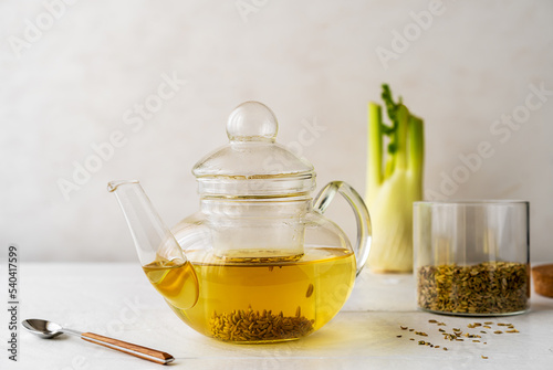 Fennel tea in a glass teapot, fresh fennel bulb, fennel seeds on white wooden table with light background
