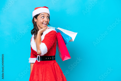 Young hispanic woman dressed as mama noel isolated on blue background holding shopping bags and smiling
