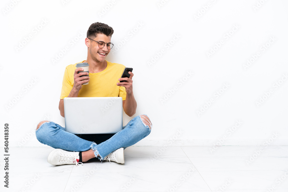 Young man sitting on the floor isolated on white background holding coffee to take away and a mobile Young man sitting on the floor isolated on white background holding coffee to take away and a mobile
