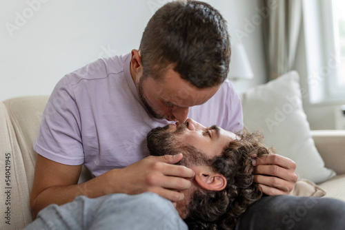 Lovely gay couple kissing while sitting in their living room at home. Two romantic young male lovers having fun indoors. Young gay couple living together.