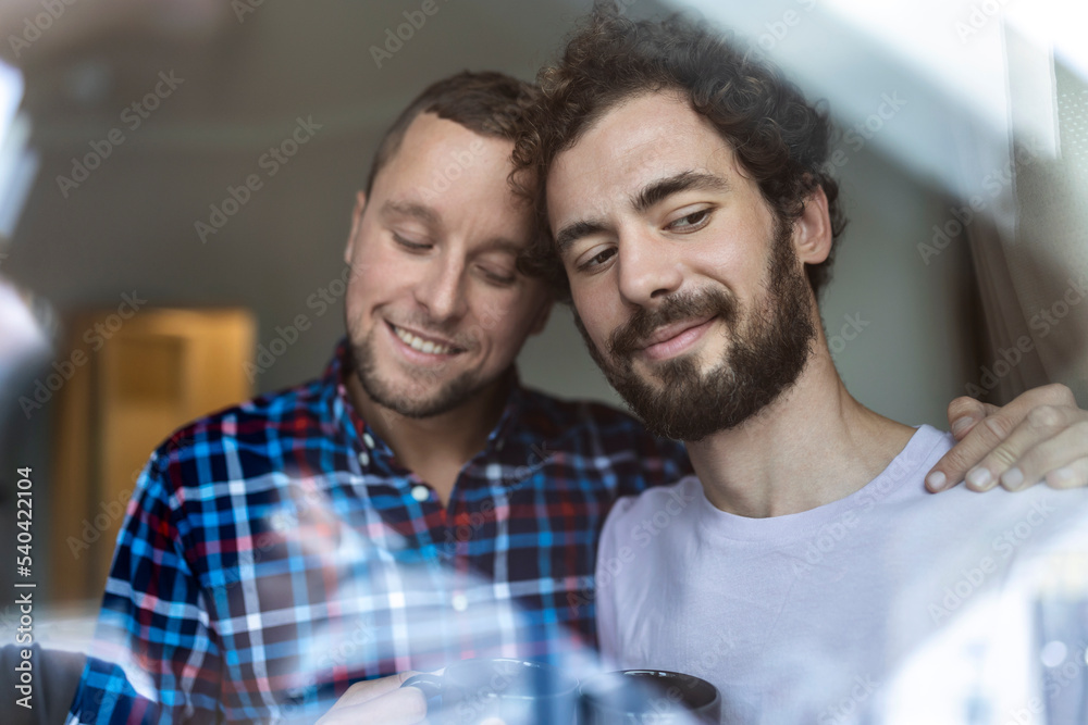 Fototapeta premium Young gay couple in love looking out the window. Two young androgynous men smiling together and having coffee.