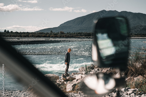 caucasian boy disheveled photographer standing looking at camera standing on a rock by the river with the van parked near mountains and nature, taramakau river, new zealand photo