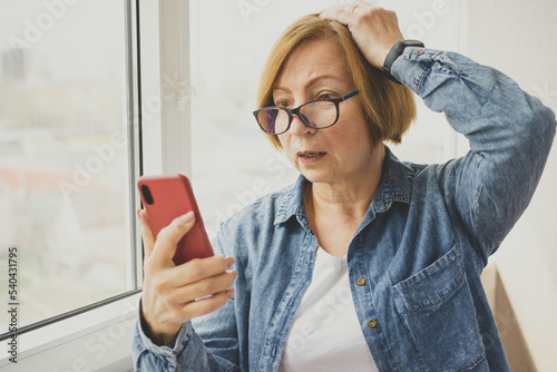 Worried mature woman looking at smartphone with amazement open mouth, reading disturbing news in media, watching horrifying video content standing near window indoor, using 5g internet connection