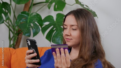 Happy young woman with credit card using instant mobile payments at home. Smiling beautiful girl shopping online, making purchase on smart phone. Monstera plant in background