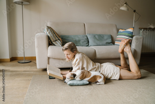 Attractive blonde woman enjoying leisure time reading novel home with small friend dog Jack Russell terrier sleeping. Relaxed moments chilling at home with your pet. Slow life atmosphere vibe