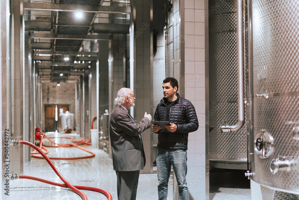 Young male vintner and senior male client walking through a winery production plant and talking about wine fermentation facts read from a touchpad