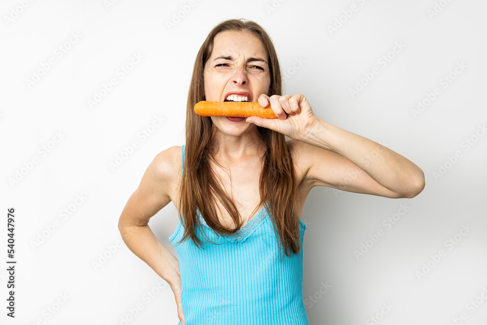Young woman screwing up her eyes biting a fresh carrot on a white background Young woman screwing up her eyes biting a fresh carrot on a white background