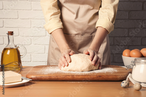 Woman kneading dough at wooden table near white brick wall, closeup