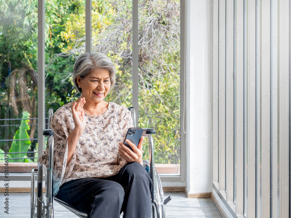 Happy Asian senior woman sits in wheelchair, waving greetings, smile and looking at smart mobile phone screen. Elderly females meet family via video call near glass window with natural green view. Happy Asian senior woman sits in wheelchair, waving greetings, smile and looking at smart mobile phone screen. Elderly females meet family via video call near glass window with natural green view.
