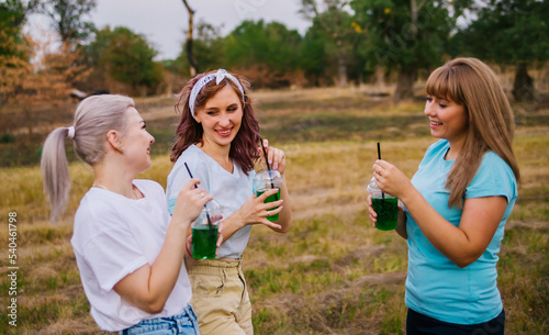 Three young women in fashionable clothes  out of town in nature  in summer and drinking refreshing non-alcoholic cocktails from disposable plastic cups. Female friendship. Holidays  pleasant leisure.