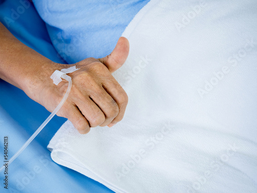 Close up saline solution in senior patients hand with thumb up in blue gown lying on the bed with white blanket in hospital room with copy space. Elder patient's hand receiving iv saline solution.