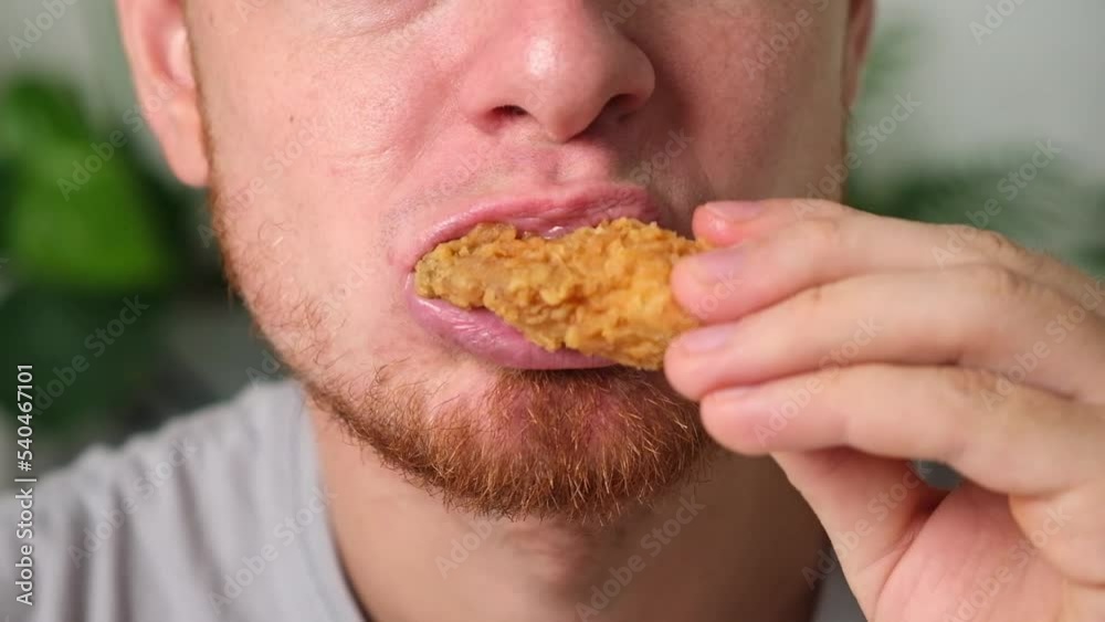 A man bites and chews a fried chicken wing in breadcrumbs. A man eats fast food. Unhealthy diet, bad habits. A man bites and chews a fried chicken wing in breadcrumbs. A man eats fast food. Unhealthy diet, bad habits.