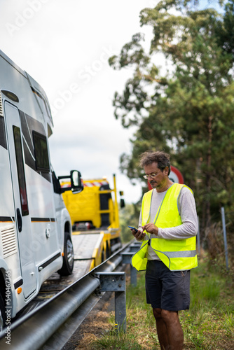 Caucasian man waiting tow truck motor home on the highway with a reflective vest and checking his phone
