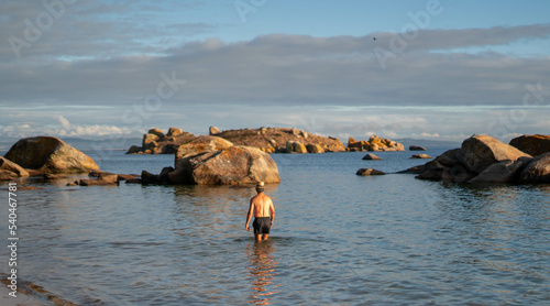 Caucasian man entering at water with a hat and blue swimsuit with rocks in the background