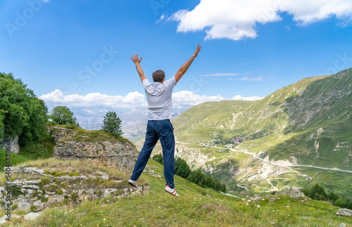 Excited millennial male traveler jumping on grassy hillside in summer mountains. Having fun, Summer vacation holiday lifestyle. Freedom and success concept.