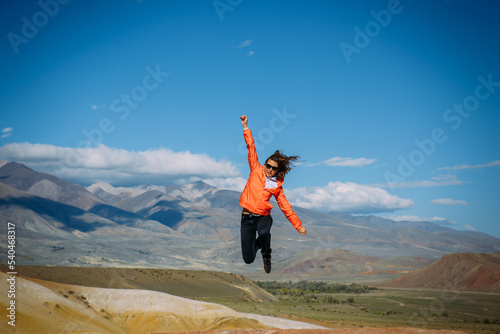 Young female traveller jumping with hands up against the mountains and blue sky. Happy woman enjoying active vacation. Travel and fun concept.