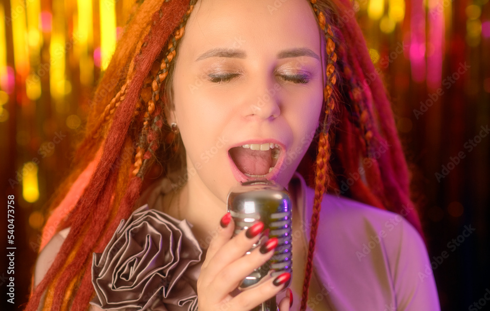Young woman with ginger dreadlocks listening to melody, singing into microphone. Portrait of female singer with closed eyes having fun in studio.