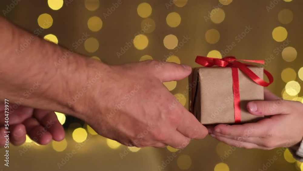 Male hands giving Christmas gift box in wrapping paper with red bow to kid girl hands. Person holding present with red ribbon on background of lights. Concept of family holidays, New Year, celebration