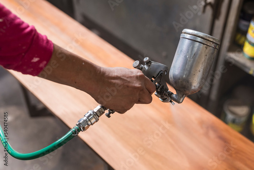 A handyman sprays lacquer wood finish on a bench or table. Using an airbrush for the application. At a furniture and woodworking shop.