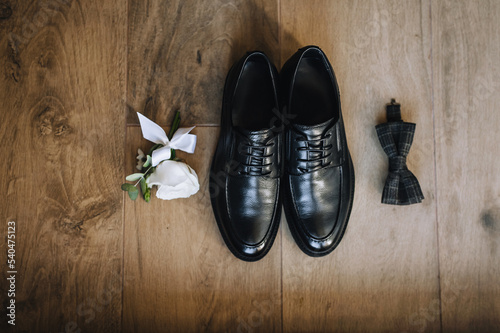 The groom's black shoes, bow tie, boutonniere lie on a wooden background.Wedding photo of accessories and details, top view.