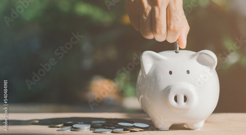 Person hand putting coins into white piggy bank in sun shine morning with copy space, coins are on wooden table. Saving money, investment, fund, loan, education, healthy, insurance, banking concept.
