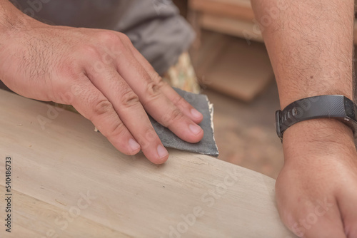 A man uses a piece of sandpaper to smooth out the edge of a circular table. Hand sanding a wood surface. A DIY project at a personal workshop.