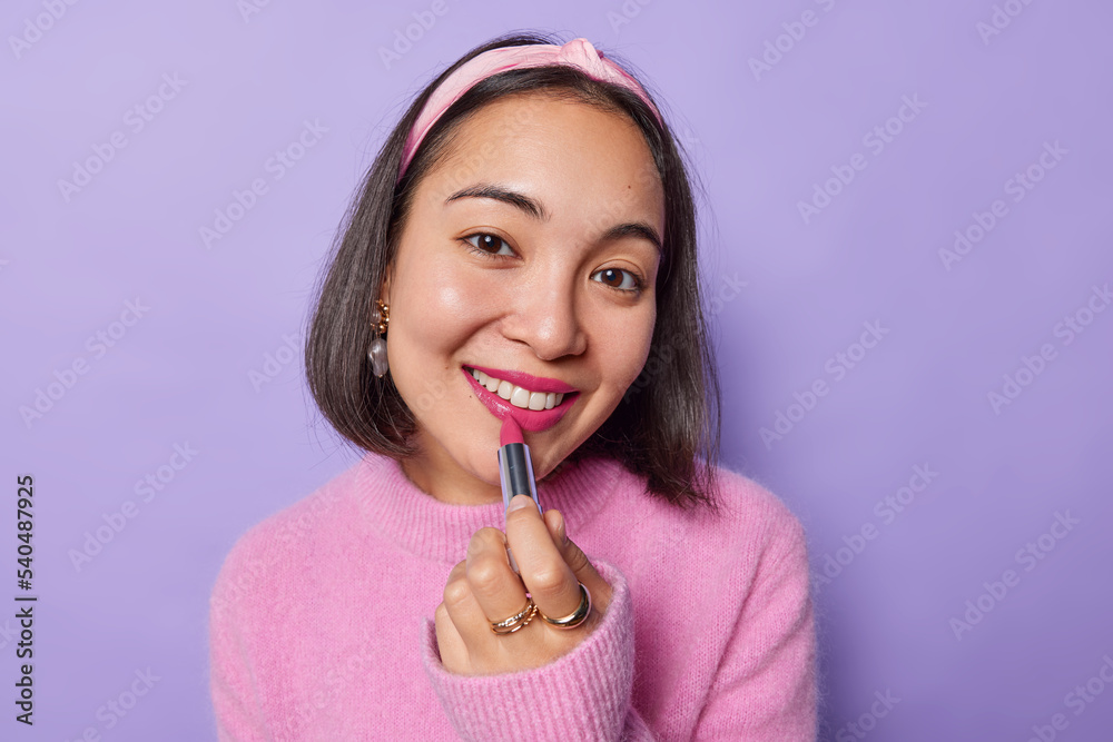 Portrait of cheerful young Asian woman with dark hair applies pink lipstick  on lips wears pink jumper and headband looks happily at camera isolated  over purple background. Daily makeup concept Stock Photo