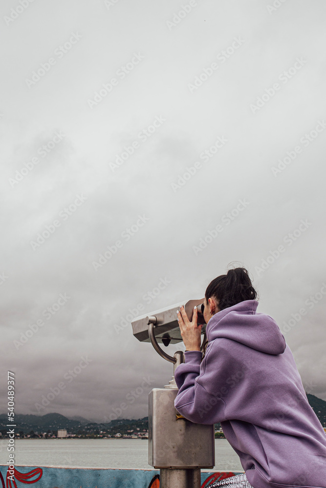 Obraz premium a girl watching through stationary binoculars on the pier for passing ships and wildlife on the seashore
